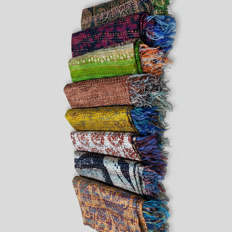 Eight colorful sari silk scarves with fringed ends are arranged in a row on a textured off-white woven rug. The scarves feature various patterns and colors including green, purple, brown, yellow, red, blue, and orange.