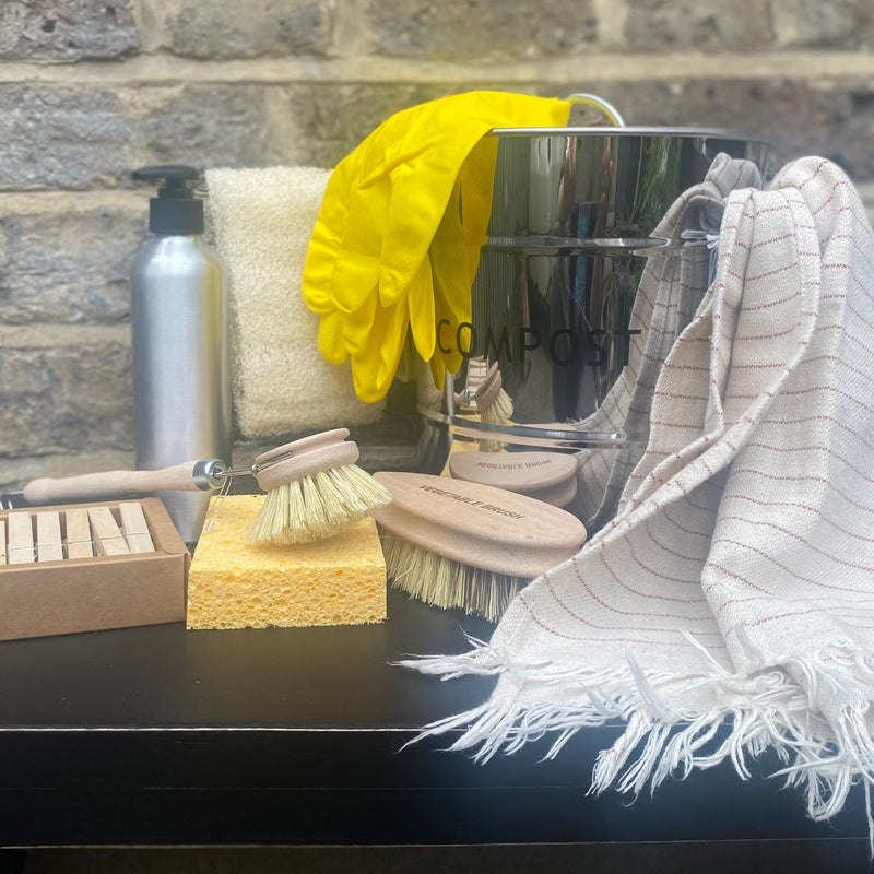 A cleaning kit arranged on a dark table against a gray brick wall, including a striped white fringed cloth, a yellow sponge, a metal bottle, a yellow glove, a metal container, and wooden-handled brushes.