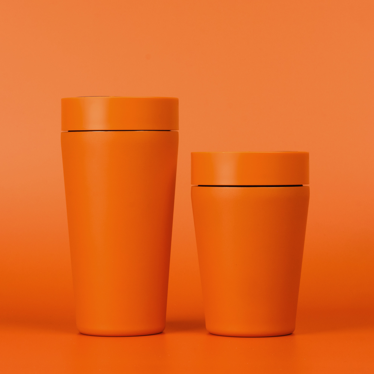 Two matte orange insulated tumblers with matching lids, one tall and one short, standing on a matching orange surface against a solid orange background.
