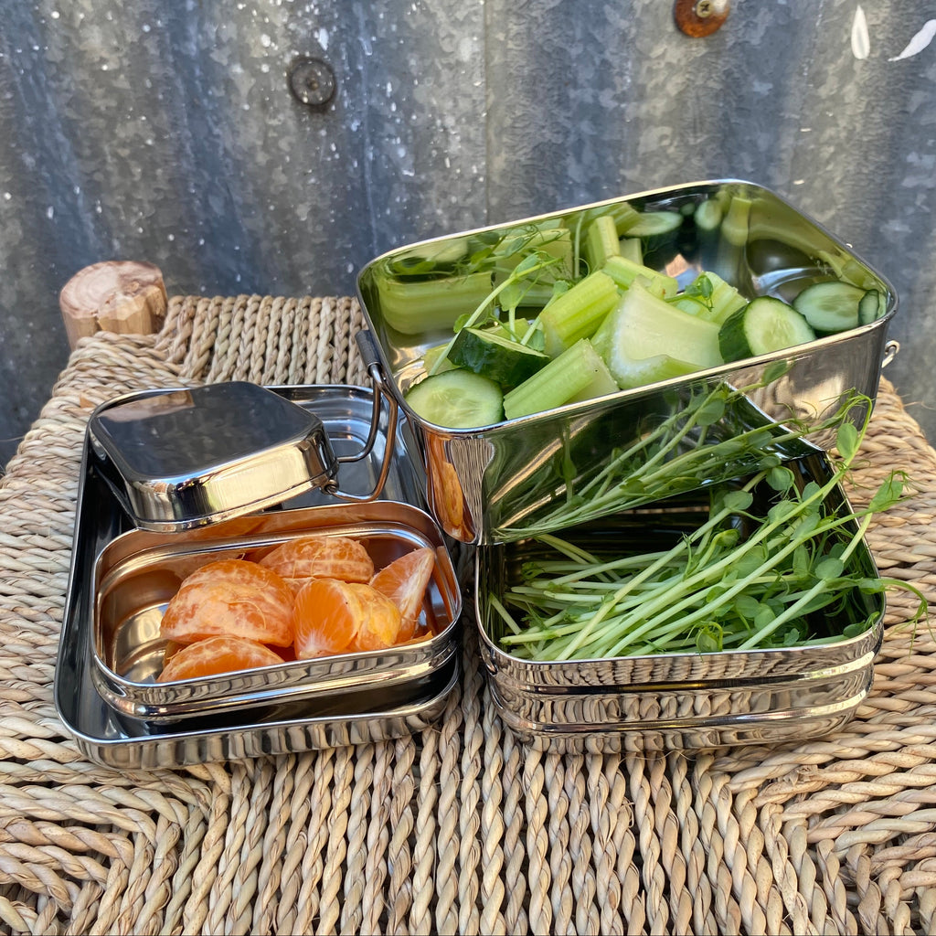 A stainless steel bento lunch box set with three  compartments, shown filled with vegetables and orange slices, placed on a woven wicker tray against a corrugated metal background.