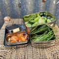 A stainless steel bento lunch box set with three  compartments, shown filled with vegetables and orange slices, placed on a woven wicker tray against a corrugated metal background.