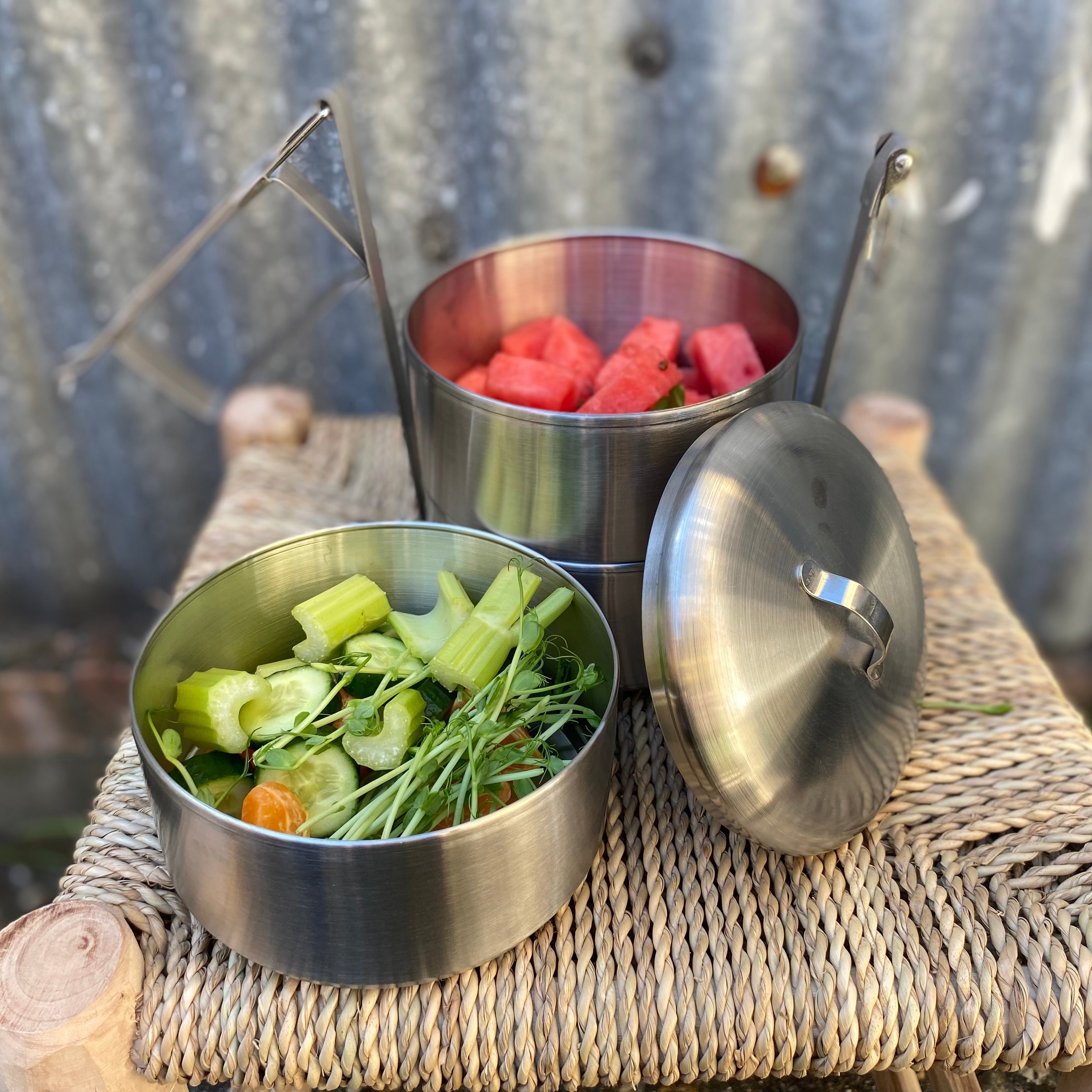 Three stainless steel bento containers with a lid, arranged on a woven straw mat outdoors. One container holds fresh green vegetables, and another holds pink watermelon chunks.