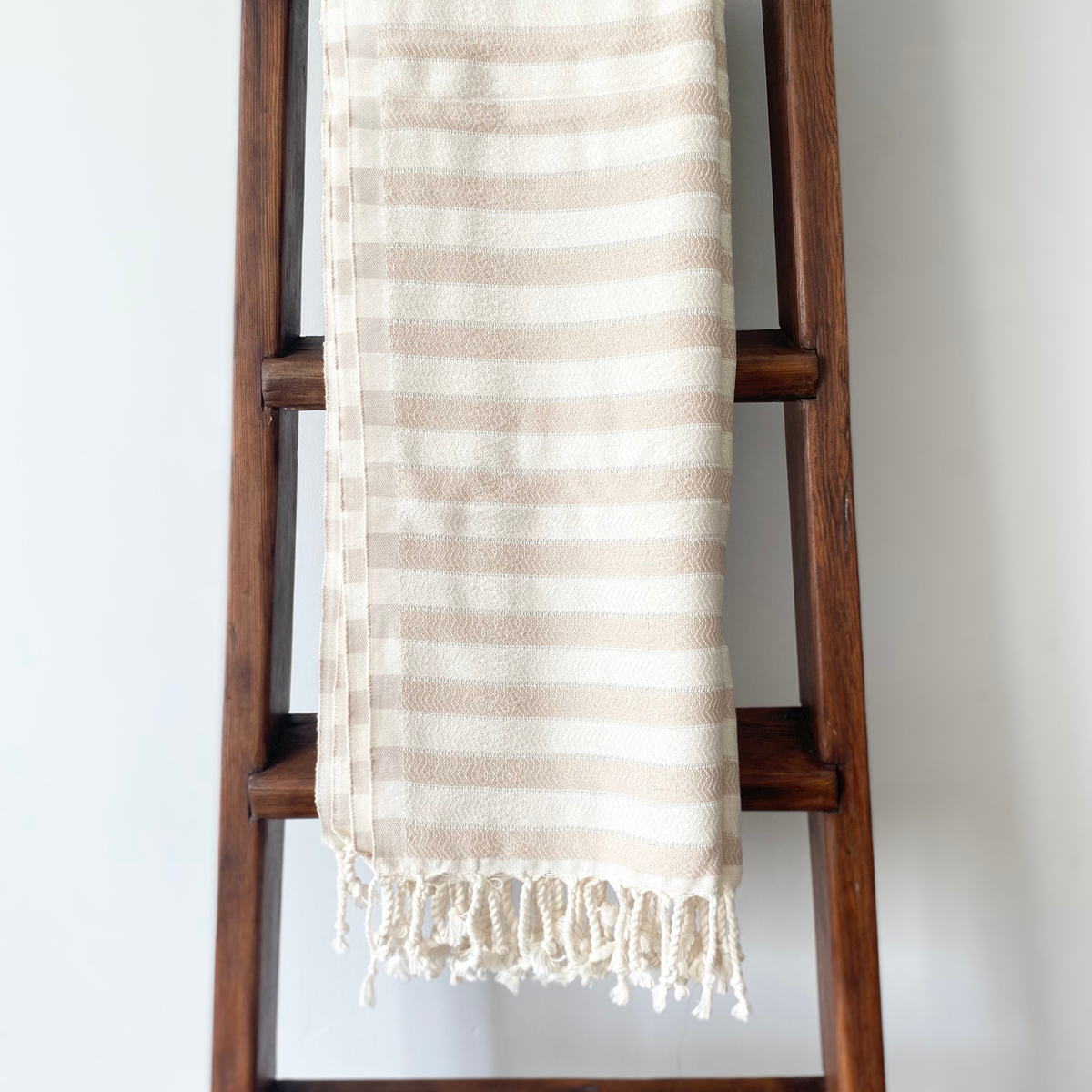 A cream and beige striped cotton Turkish towel with fringed edges draped over a dark wooden ladder against a plain white wall.