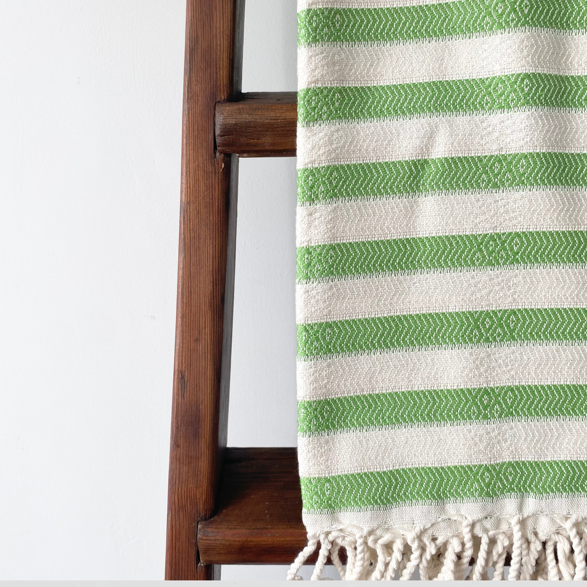 A close-up of a woven Turkish cotton towel with green and off-white stripes, draped over a dark wooden ladder against a plain white wall.