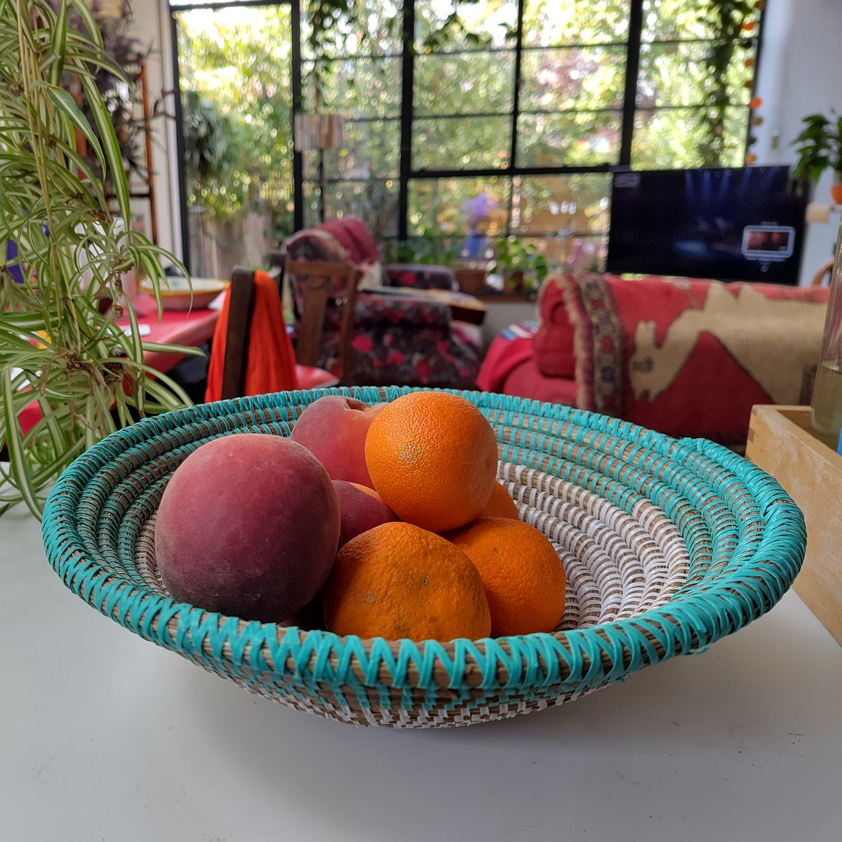 A round woven fruit basket with a turquoise and white pattern sits on a white table in a bright, modern living room. Inside the basket are several oranges and a pinkish fruit.