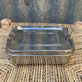 A rectangular stainless steel lunch box with a fitted lid and side clips, placed on a woven straw or wicker surface against a corrugated metal wall.