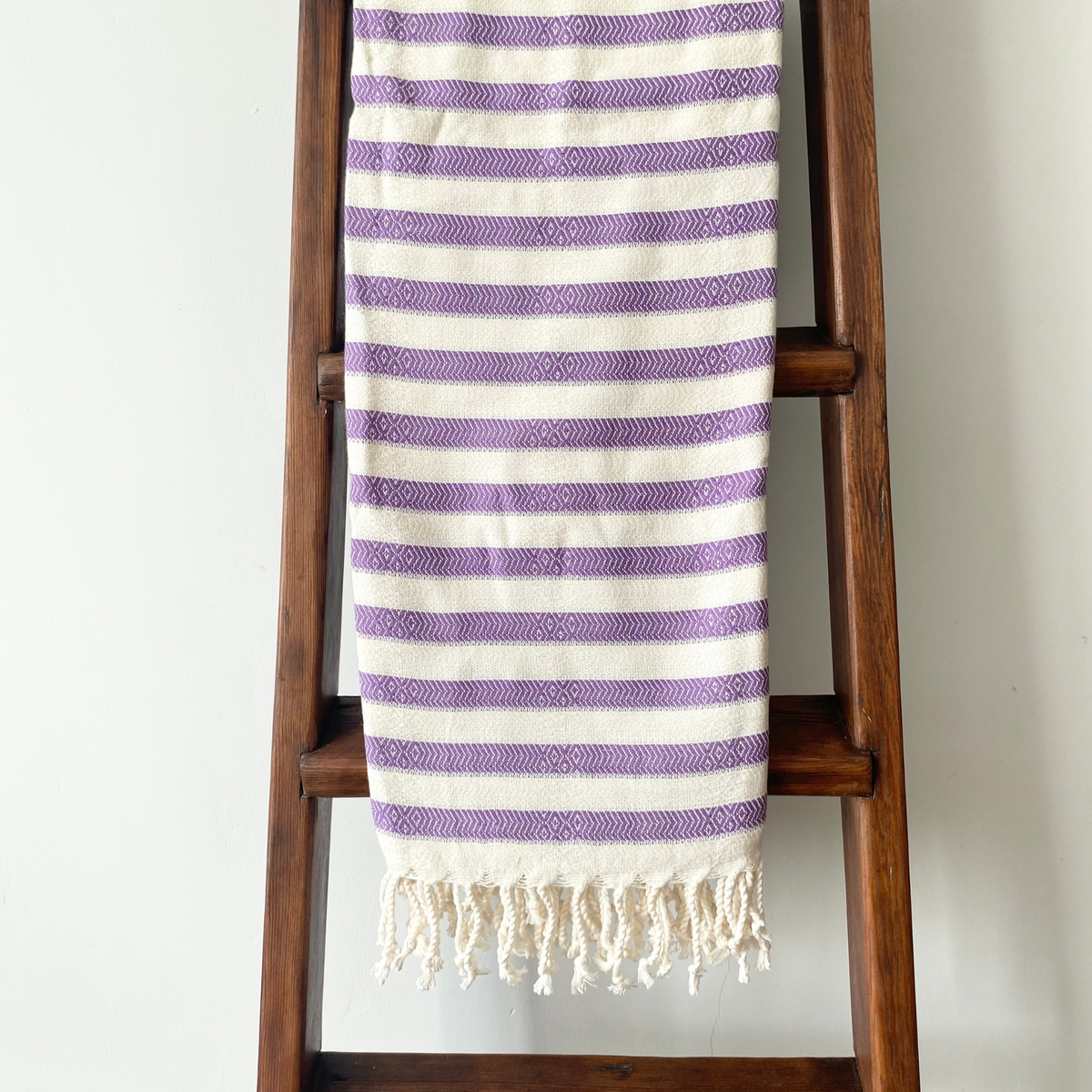 A striped woven Turkish towel with cream and purple horizontal stripes and fringed tassels, draped over a dark wooden ladder against a plain white wall.