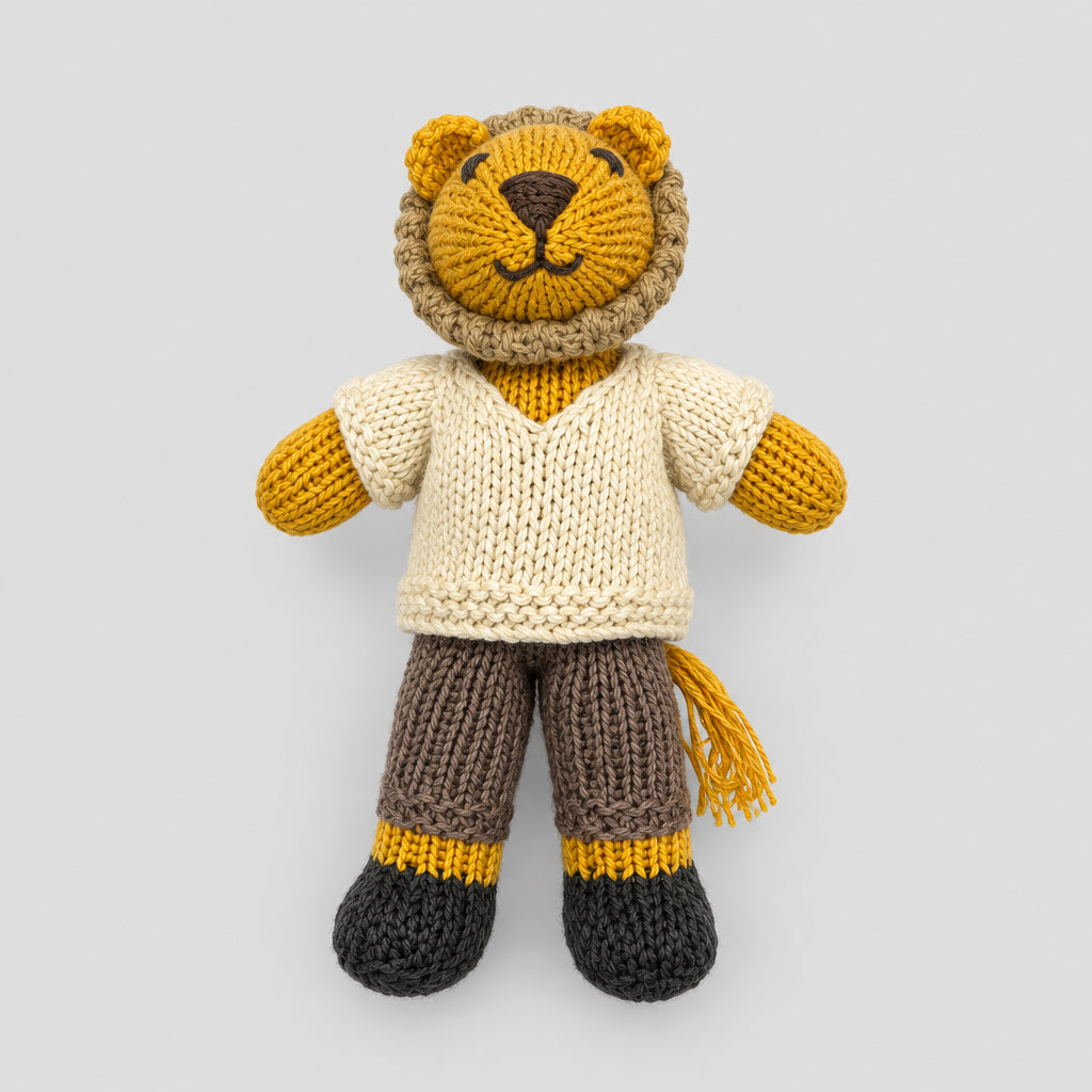 A handmade knitted lion doll wearing a cream sweater, brown trousers, and dark grey boots, lying on a white fabric background.
