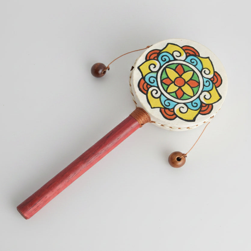 A traditional hand drum, featuring a hand-painted circular head drum with a yellow, red, blue, and green mandala pattern, and a reddish-brown wooden handle with two hanging wooden beaters.