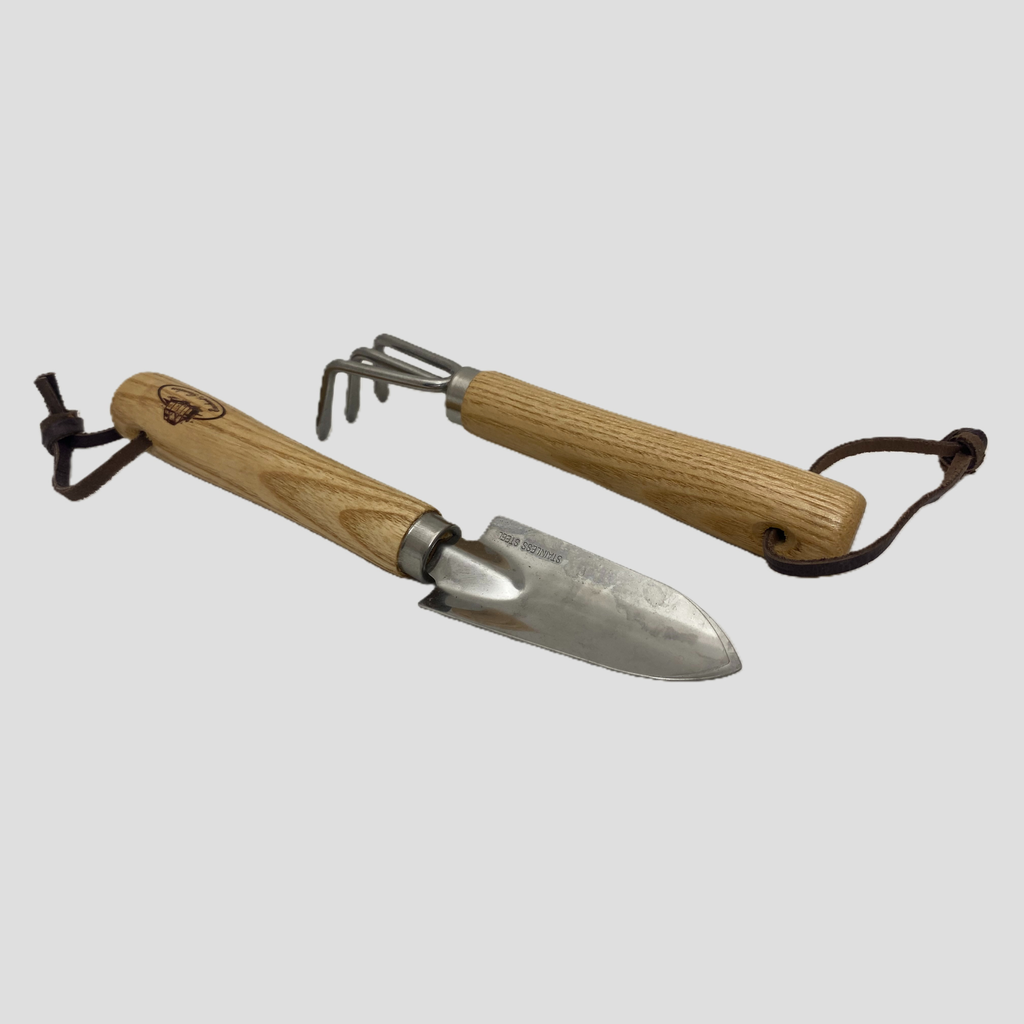 A small hand garden tool set with two wooden-handled tools on a light gray background. One tool is a trowel with a steel blade and a dark brown leather loop, and the other is a small rake with a steel head and handle, also with a leather loop.