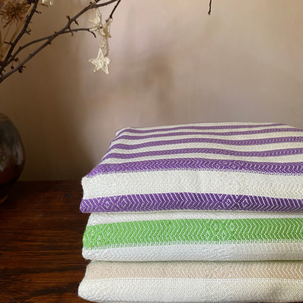 Three folded woven textiles stacked on a dark wooden surface, featuring purple and white stripes, a green and white striped band, and a beige and white striped band, with a neutral wall and dried branches in the background.