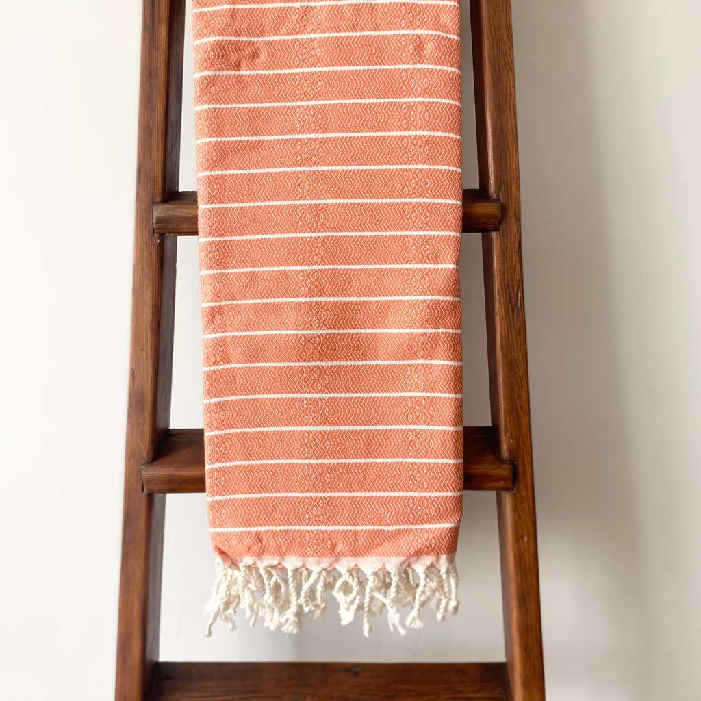 A terracotta orange and white striped Turkish cotton towel with fringed edges, draped over a dark brown wooden ladder against a plain off-white wall.