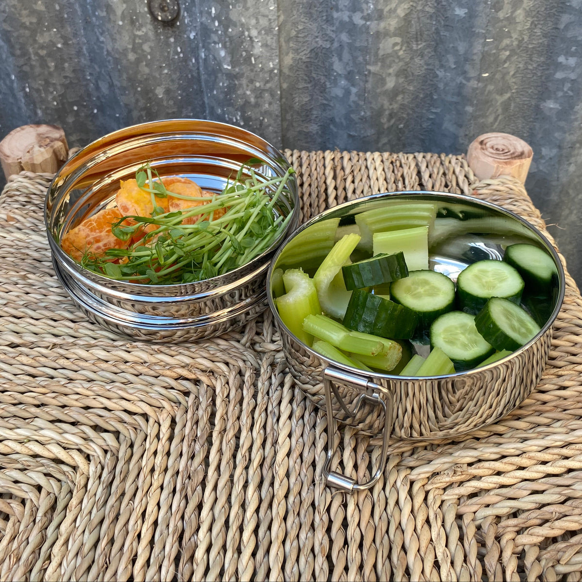 Two round stainless steel lidded containers placed on a woven straw chair outdoors, with one container open and filled with sliced cucumbers in water and the other showing cooked food inside.