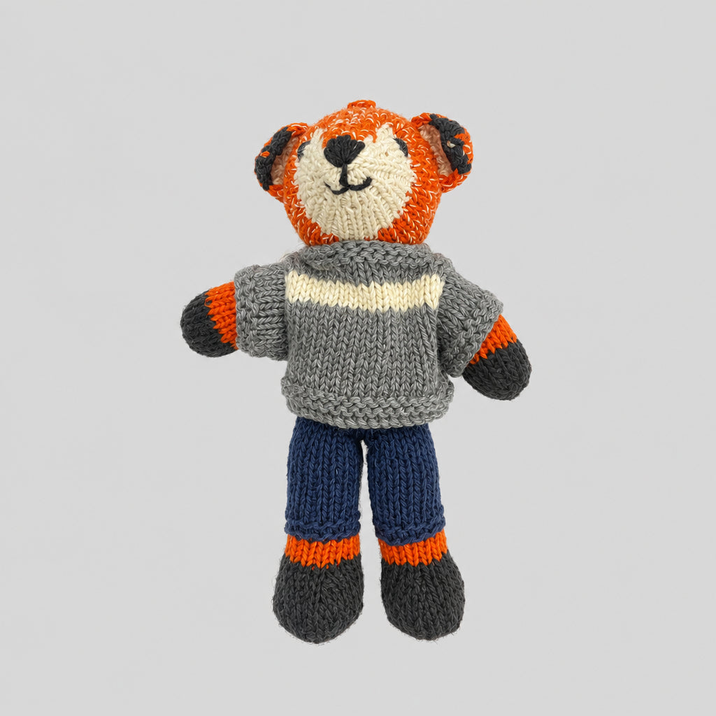 A hand-knit teddy fox doll wearing a green sweater with cream stripes, blue pants, and orange and gray accents.
