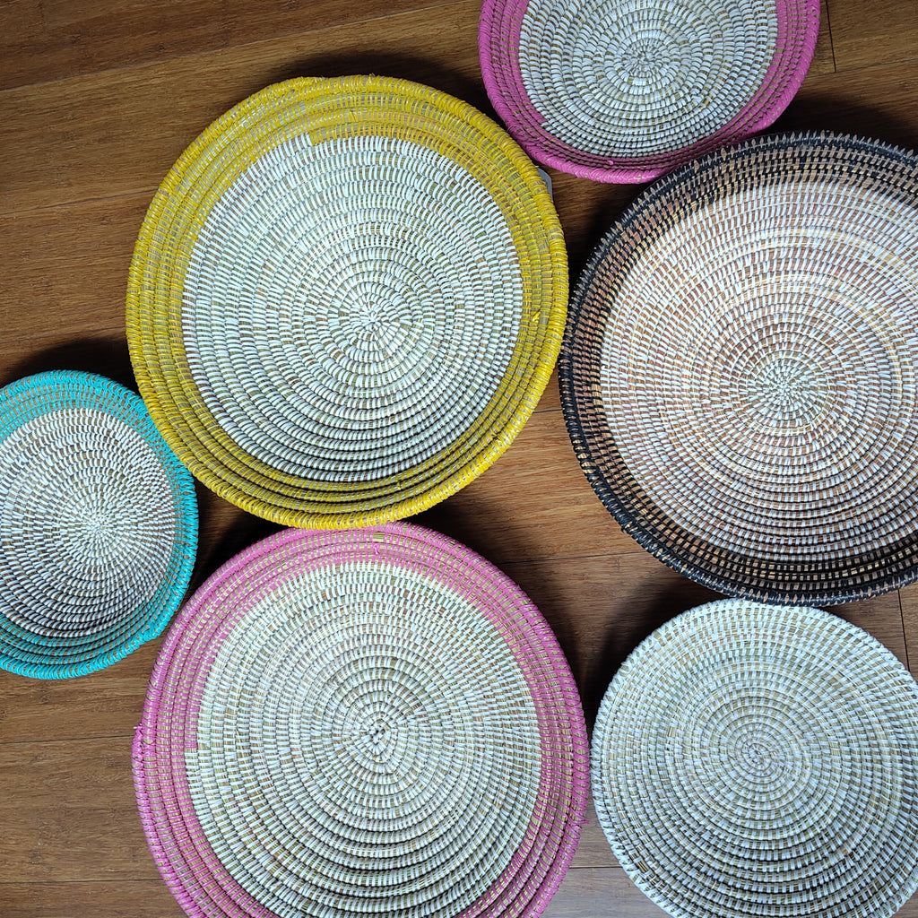 Six round woven basket bowls in various colors and sizes arranged on a wooden floor. The bowls feature a spiral woven pattern with rims in pink, yellow, teal, black, and white, set against a warm brown wood background.