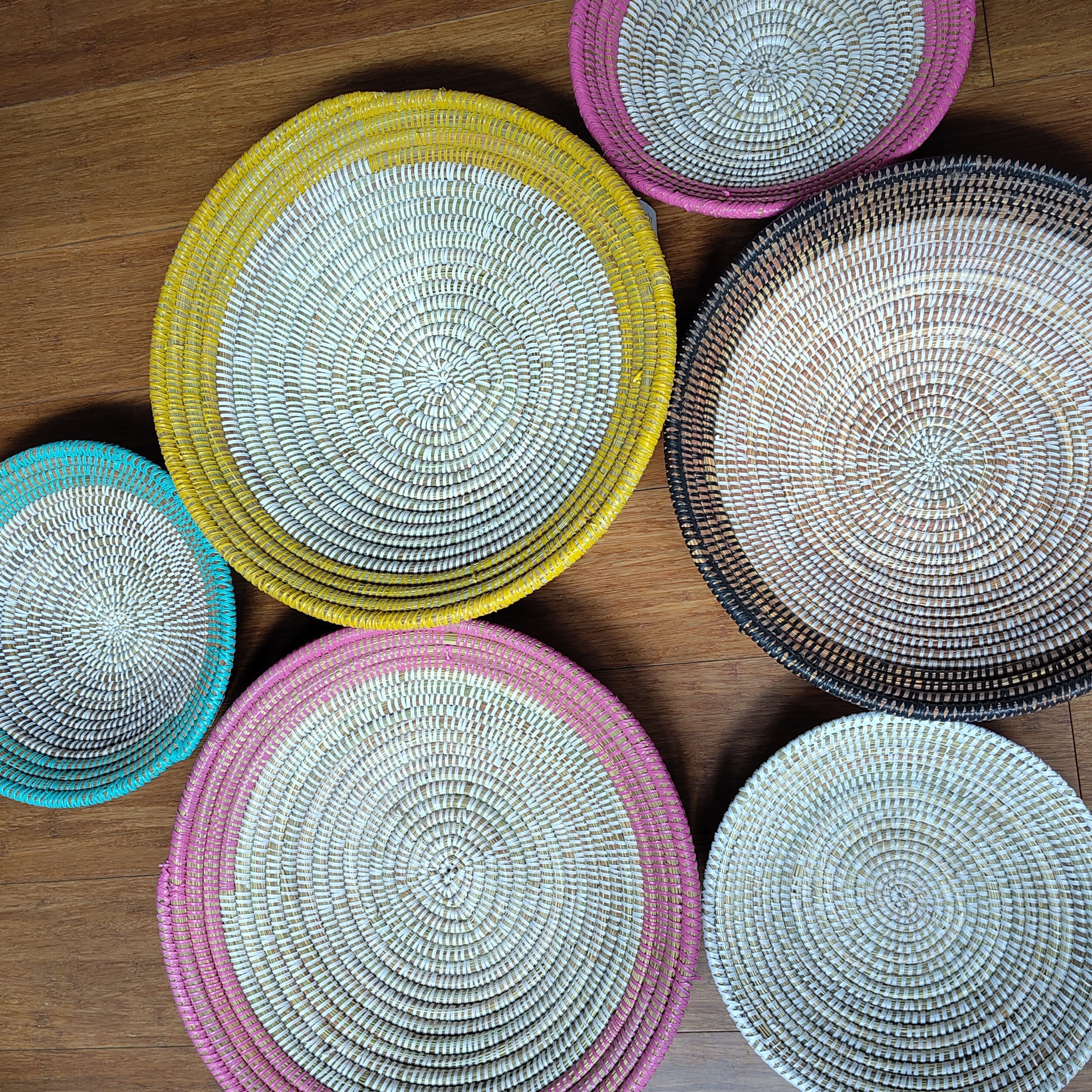 Six round woven basket bowls in various colors and sizes arranged on a wooden floor. The bowls feature a spiral woven pattern with rims in pink, yellow, teal, black, and white, set against a warm brown wood background.