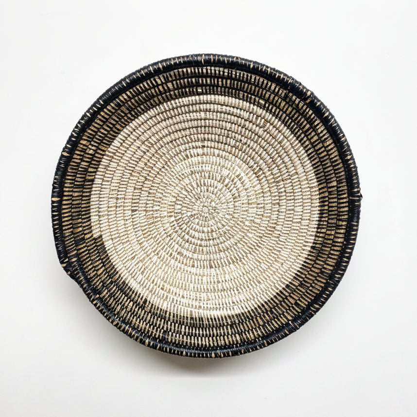 A round woven basket with a hand-woven pattern in black, beige, and brown, photographed from directly above on a white background 