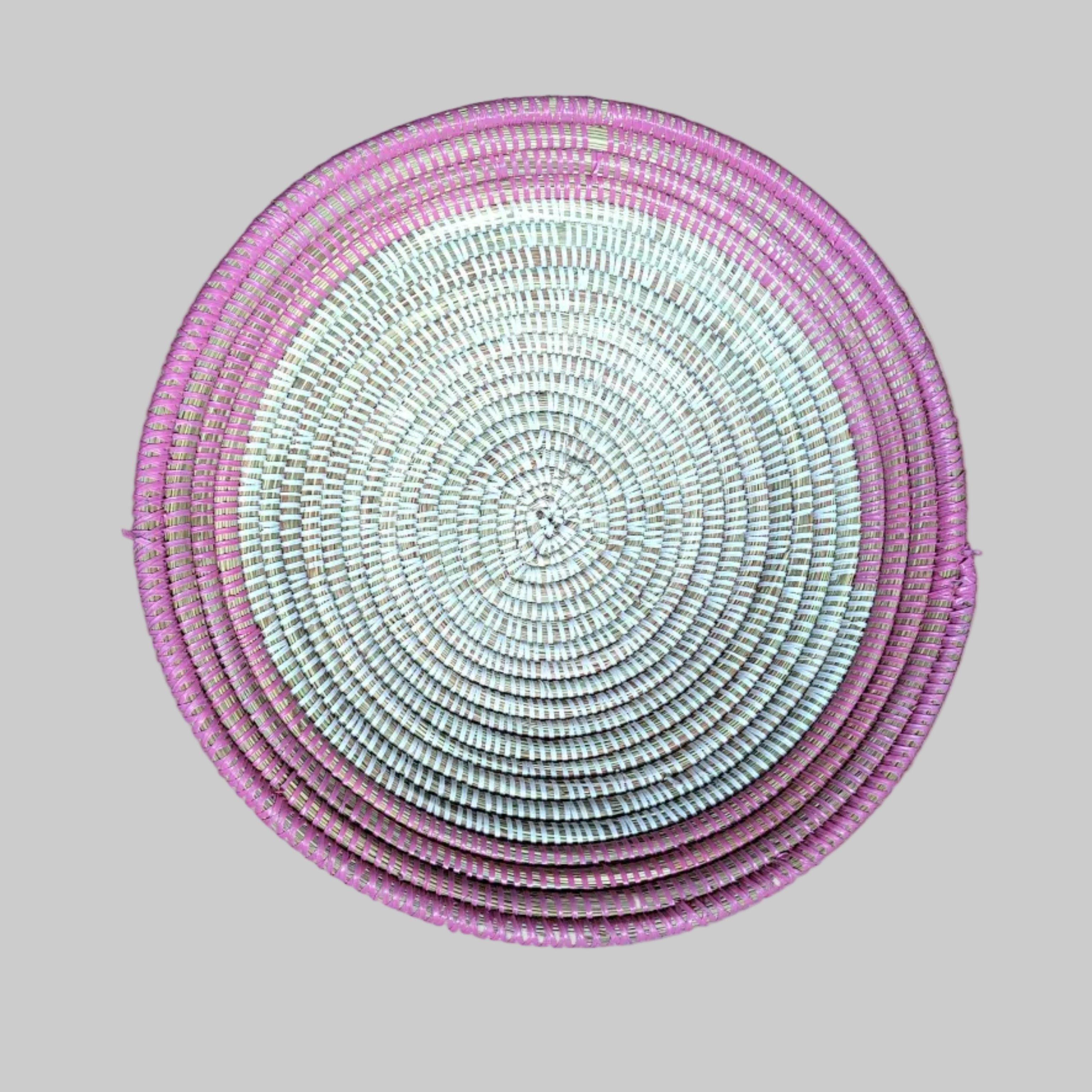 A round woven basket or tray with a pink and white spiral pattern, photographed from directly above.on a brown wooden floor.