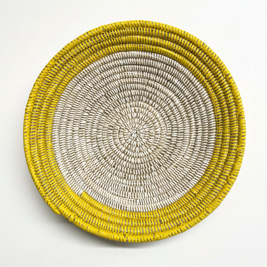 A round woven basket or tray with concentric rings, featuring a bright yellow outer rim and a natural beige woven center, photographed from directly above on a white background.