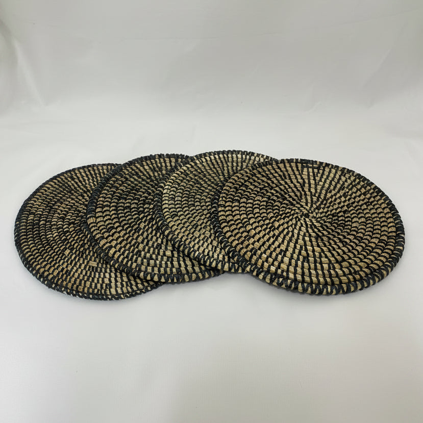 Four round woven seagrass placemats with a black and natural beige spiral pattern, arranged in a fan shape on a white fabric background.