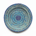 A round woven basket plate with concentric spiral patterns in blue, turquoise, and brown, displayed on a plain white background. 