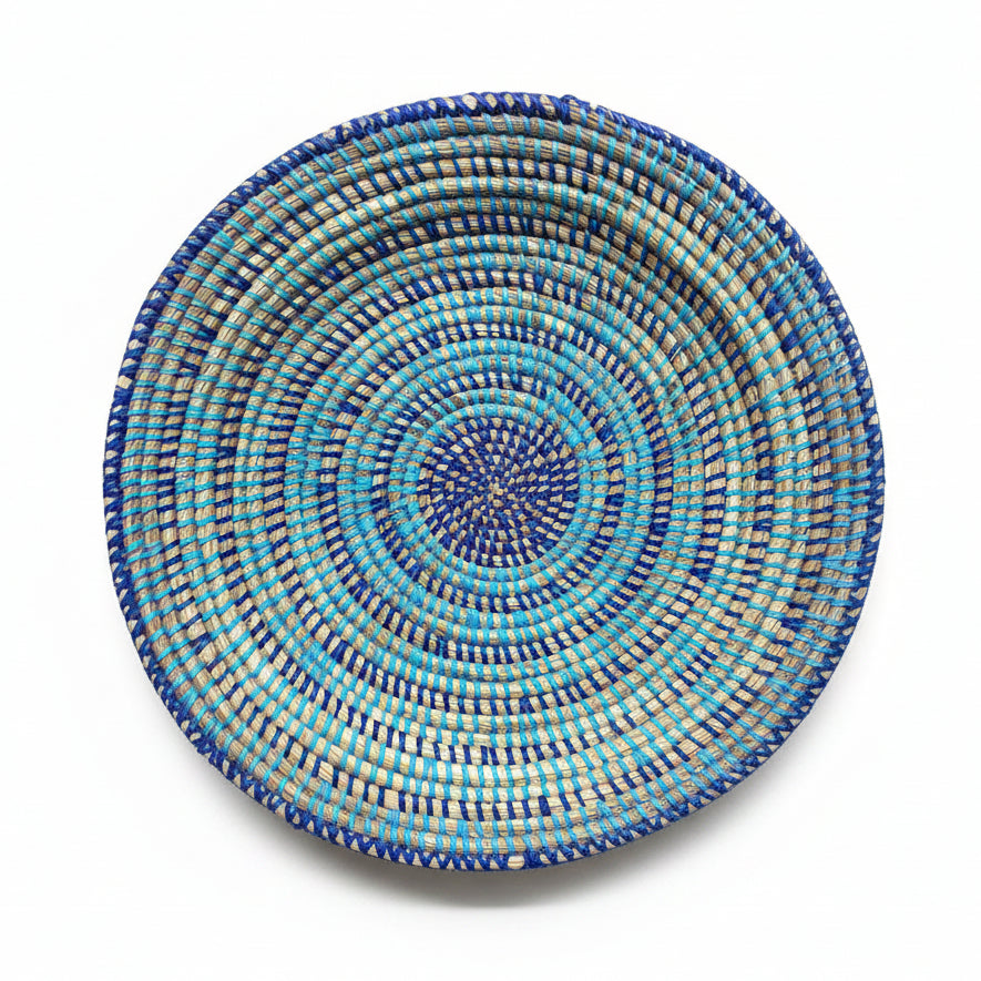 A round woven basket plate with concentric spiral patterns in blue, turquoise, and brown, displayed on a plain white background. 