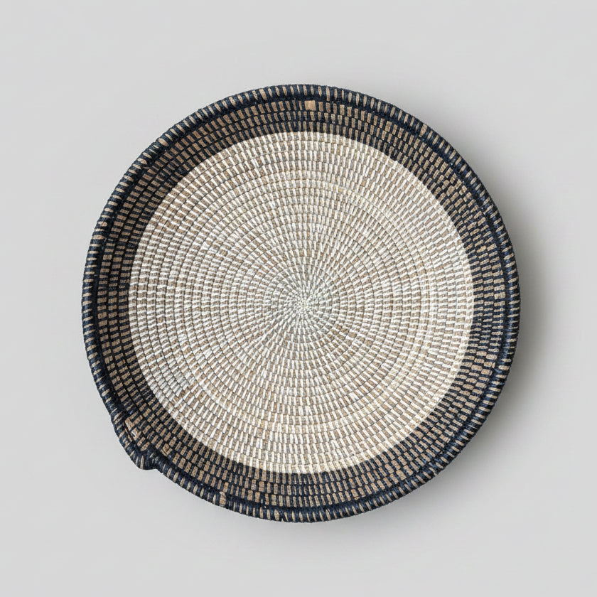 A round woven basket tray with a dark charcoal outer rim and a light beige and brown coiled center.