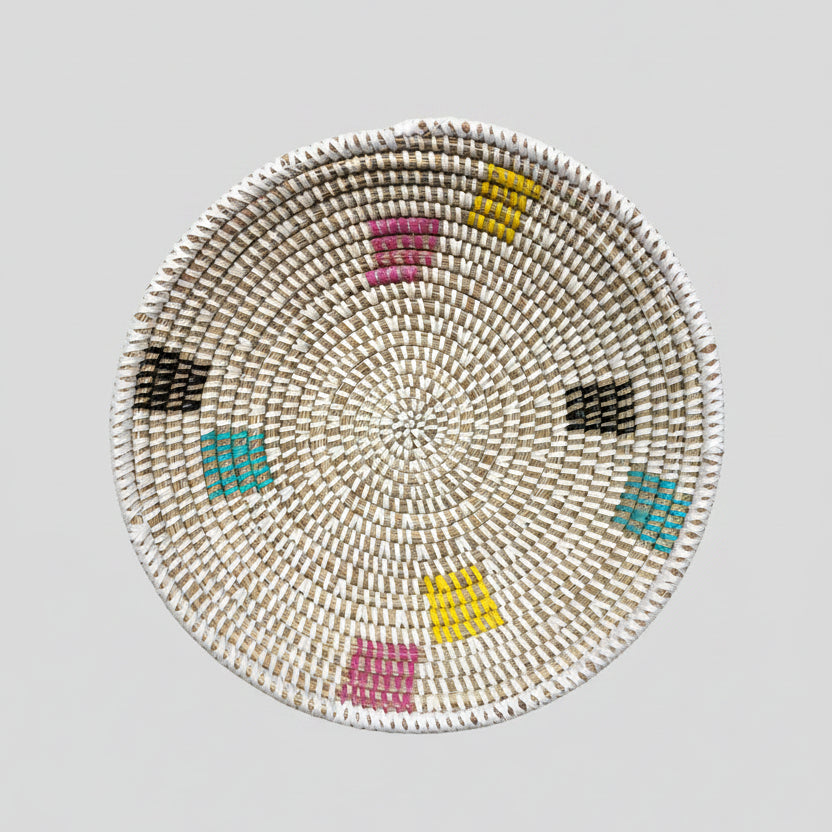 A round woven basket  with a geometric pattern in pink, yellow, turquoise, black, and white, displayed on a white background.