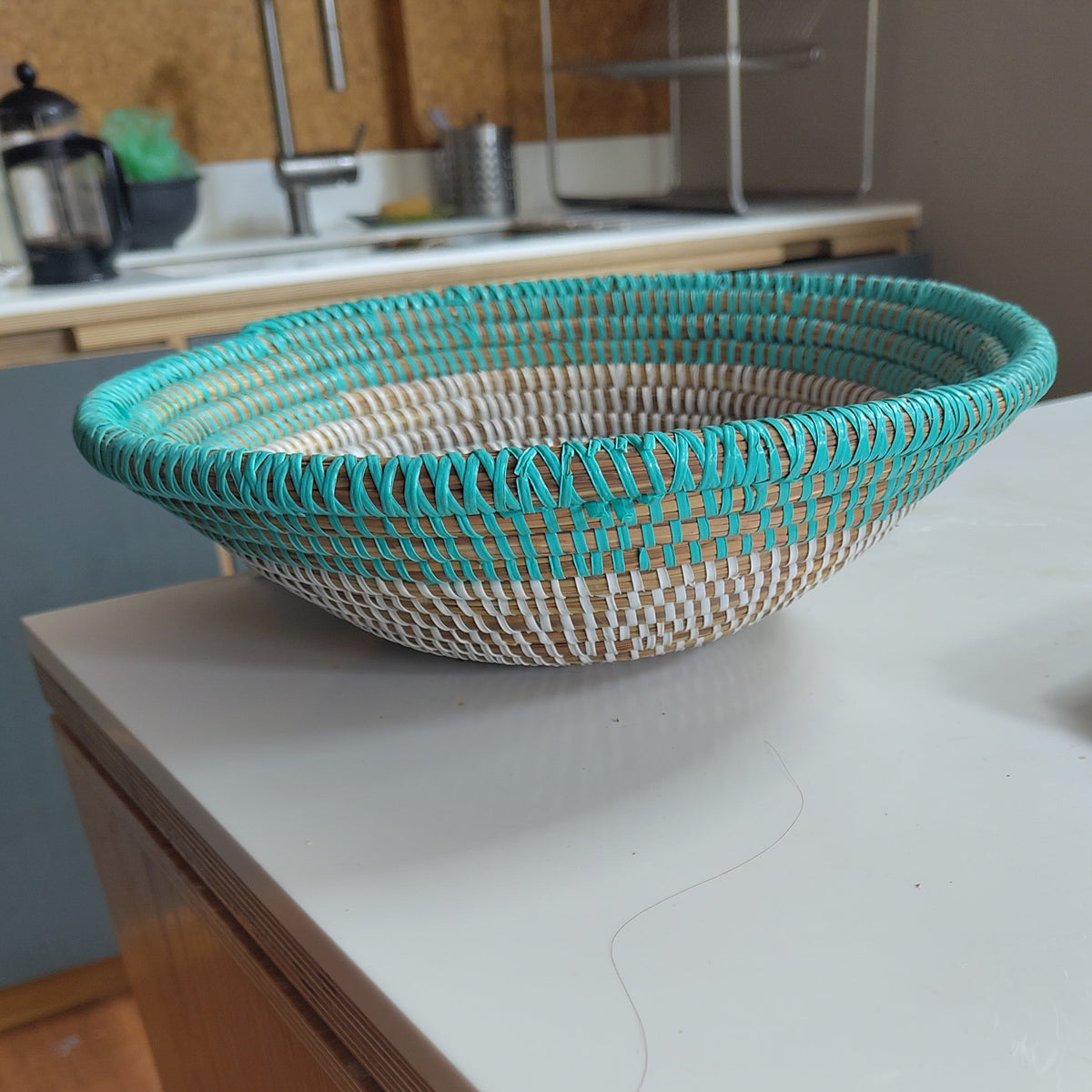 A woven oval basket with turquoise blue and natural beige stripes sits on a white kitchen countertop.