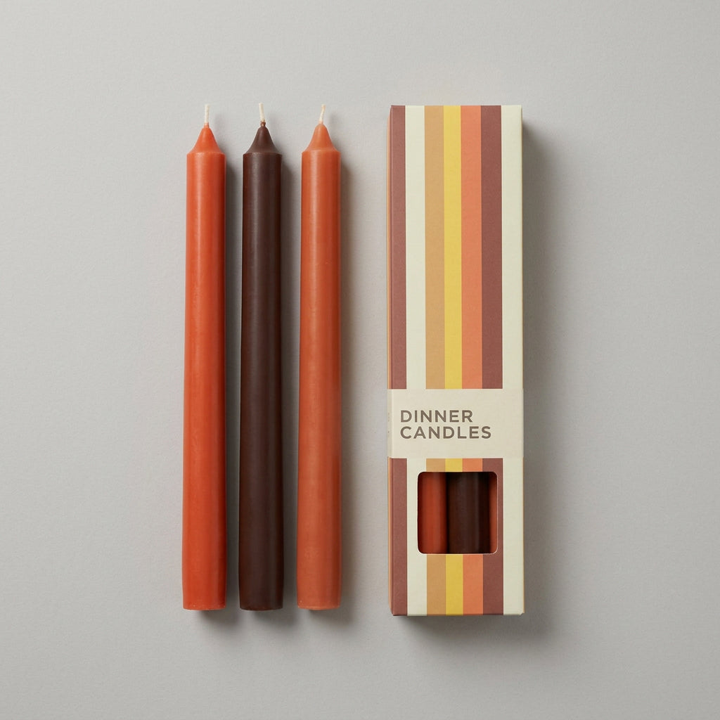 Three dinner candles, Orange, red and  dark brown, shown next to their striped cardboard packaging box.