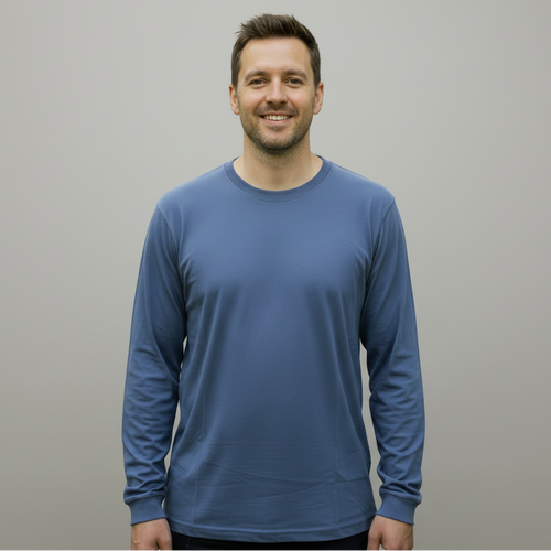 A man wearing a dusty blue long-sleeve t-shirt and dark jeans, standing in front of woodland.