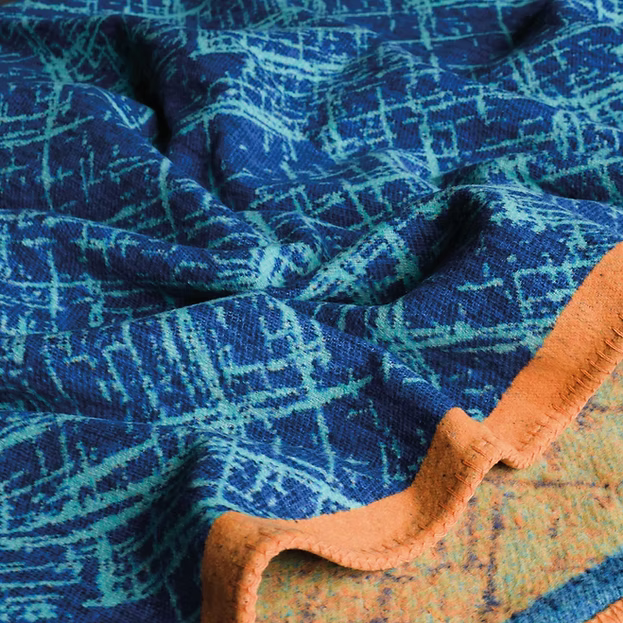 A close-up of a textured woven blanket or throw in deep blue and turquoise with a geometric pattern, featuring a contrasting orange stitched border.