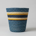 A hand-woven basket with horizontal stripes of blue, navy, mustard yellow, and cream, sitting on a rustic wooden table against a white wall.