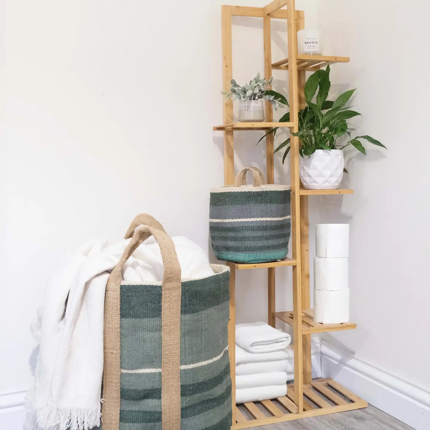 A tall light wood shelving unit styled with plants, a striped green and beige storage basket, rolled white towels, and a white throw blanket draped over a large matching woven basket on the floor. The scene is set against a plain white wall and light wood flooring.
