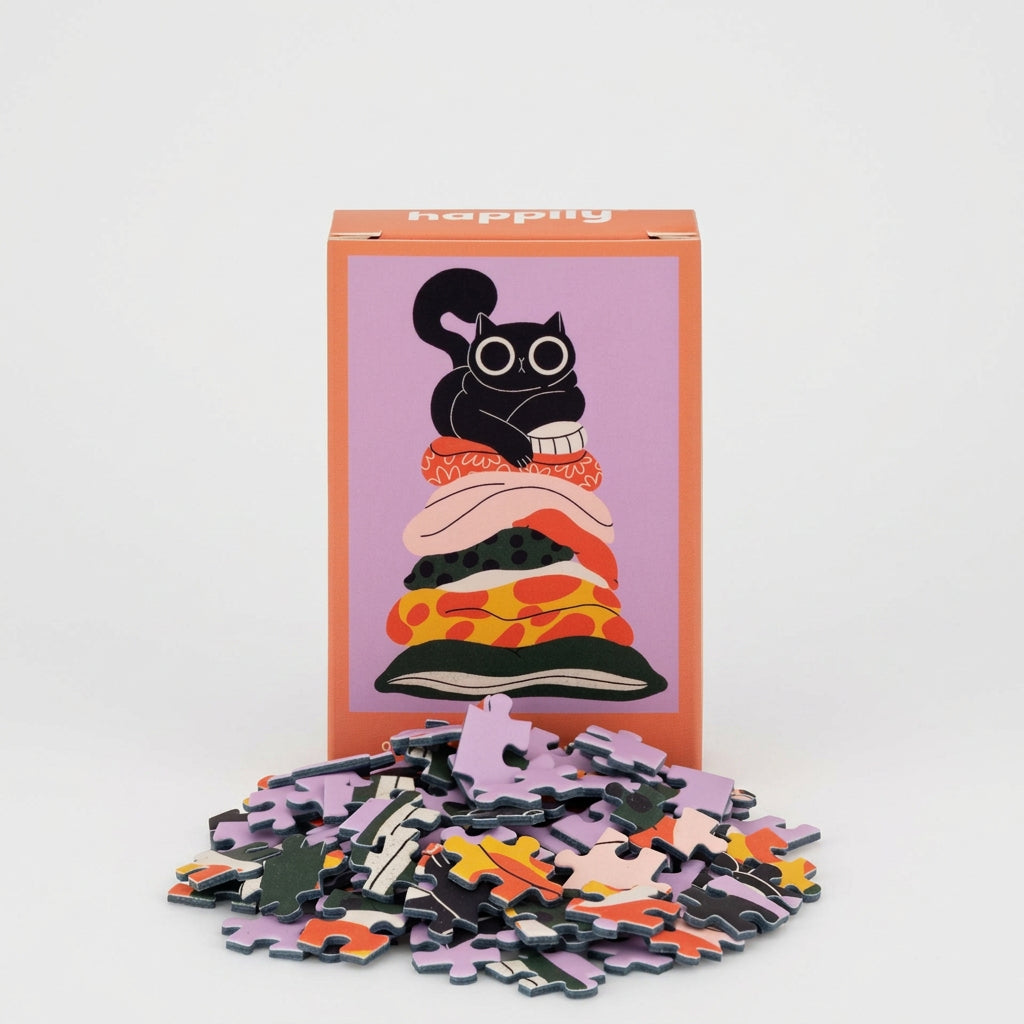 A  jigsaw puzzle box with a colorful illustration of a black cat on  cushions, shown with a pile of loose puzzle pieces in front on a grey background.