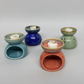 Six small ceramic wax melts or scented candle holders in assorted colors, arranged on a patterned fabric surface against a plain white wall.