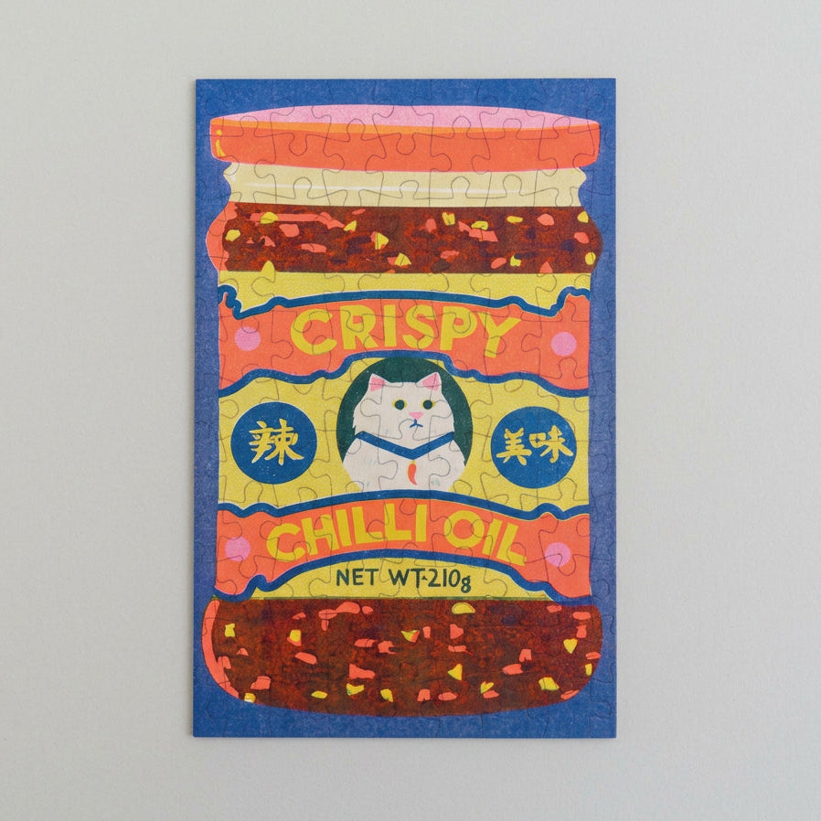 A colorful jigsaw puzzle featuring an illustrated jar of 'Crispy Chilli Oil' with a white cat on the label, set against a textured blue background.