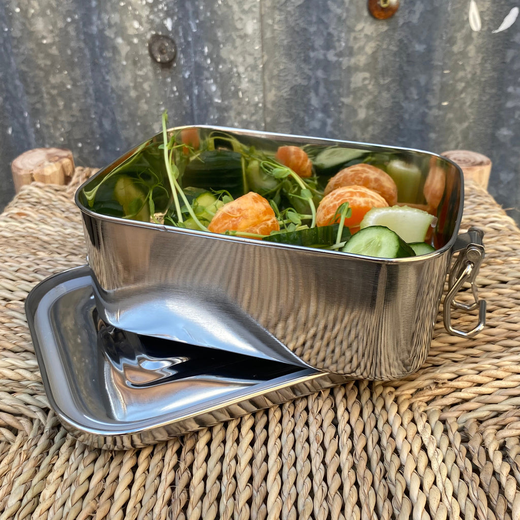 A stainless steel lunch box with a matching lid sits on a woven straw table mat outdoors, filled with a salad containing cucumber, spinach, and orange slices.