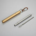 A gold colour metal folding straw caseshown on a grey surface, including   a carabiner clip and  a long metal straw and a small brush.