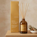 An amber glass reed diffuser bottle with wooden reeds and a gold label, standing in front of matching gold product box on a shelf.