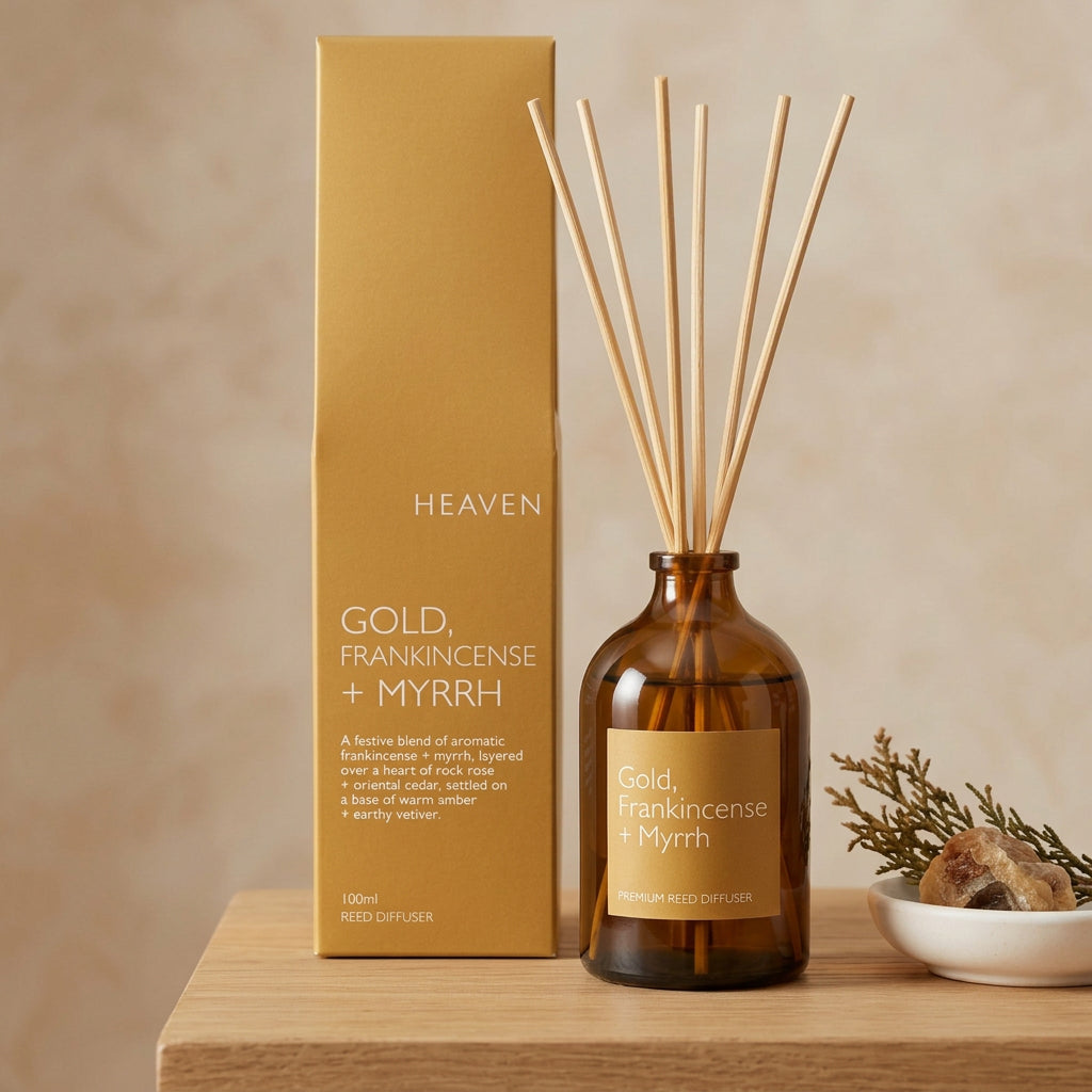 An amber glass reed diffuser bottle with wooden reeds and a gold label, standing in front of matching gold product box on a shelf.