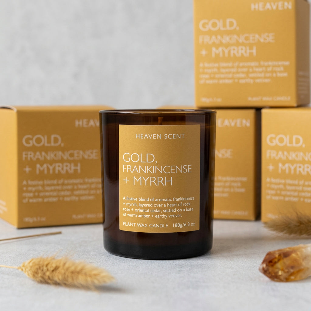 Amber glass candle jar with a mustard-yellow label reading 'GOLD, FRANKINCENSE + MYRRH' by Heaven Scent, displayed on a shelf with matching product boxes behind it.