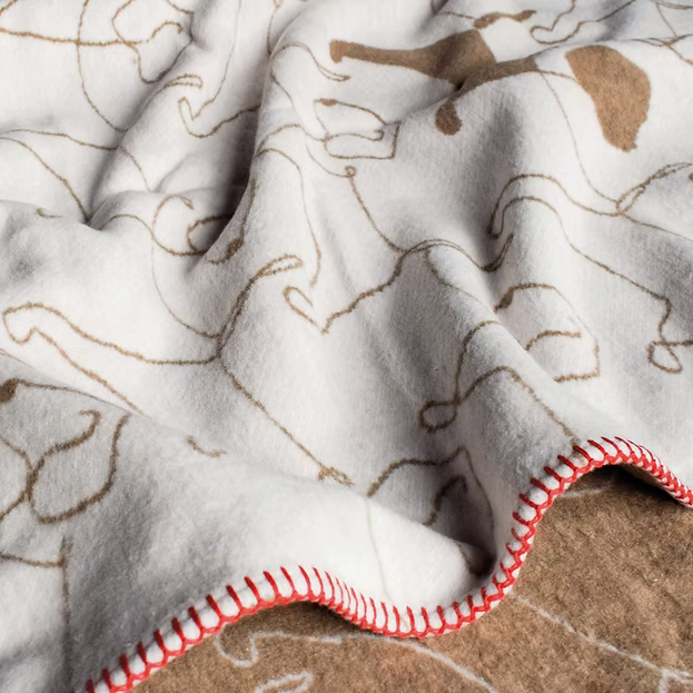 A close-up of a soft fleece blanket with a white background, brown line-art dog illustrations, and a red-and-white stitched border, reversable with a brown reverse.