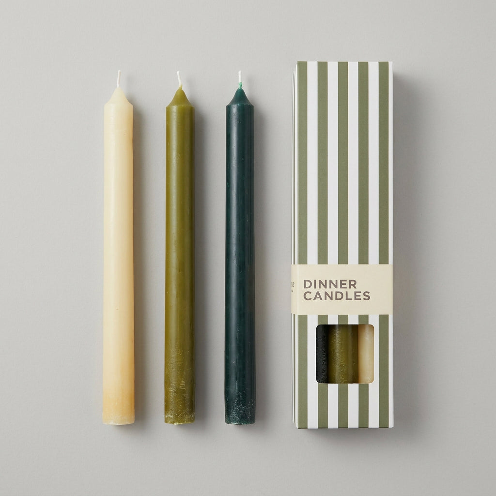 Three taper candles in cream, olive green, and dark teal are displayed next to their striped packaging box labeled 'DINNER CANDLES' .