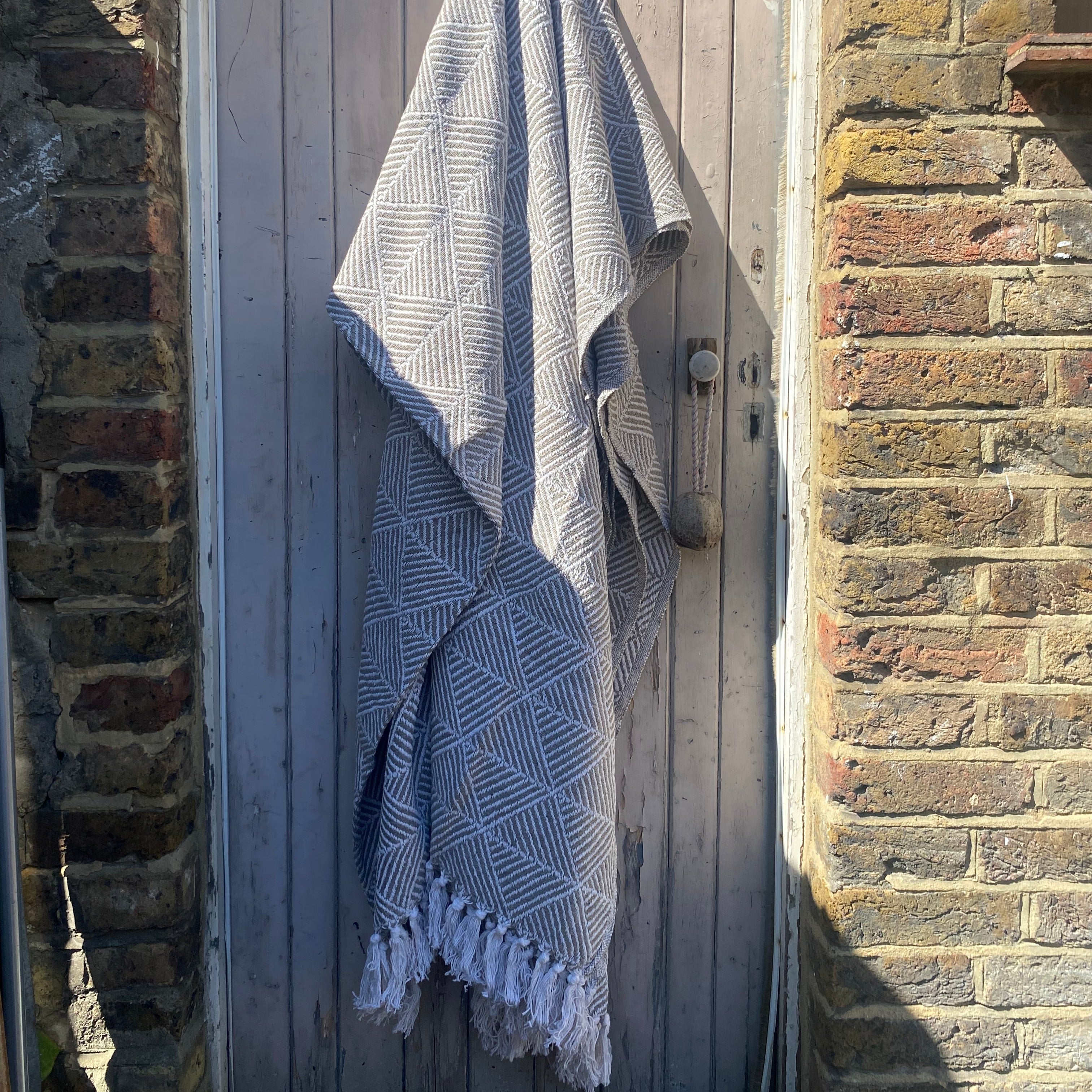 A gray and white geometric patterned throw blanket with fringed edges hanging on a weathered gray wooden door between brick walls.