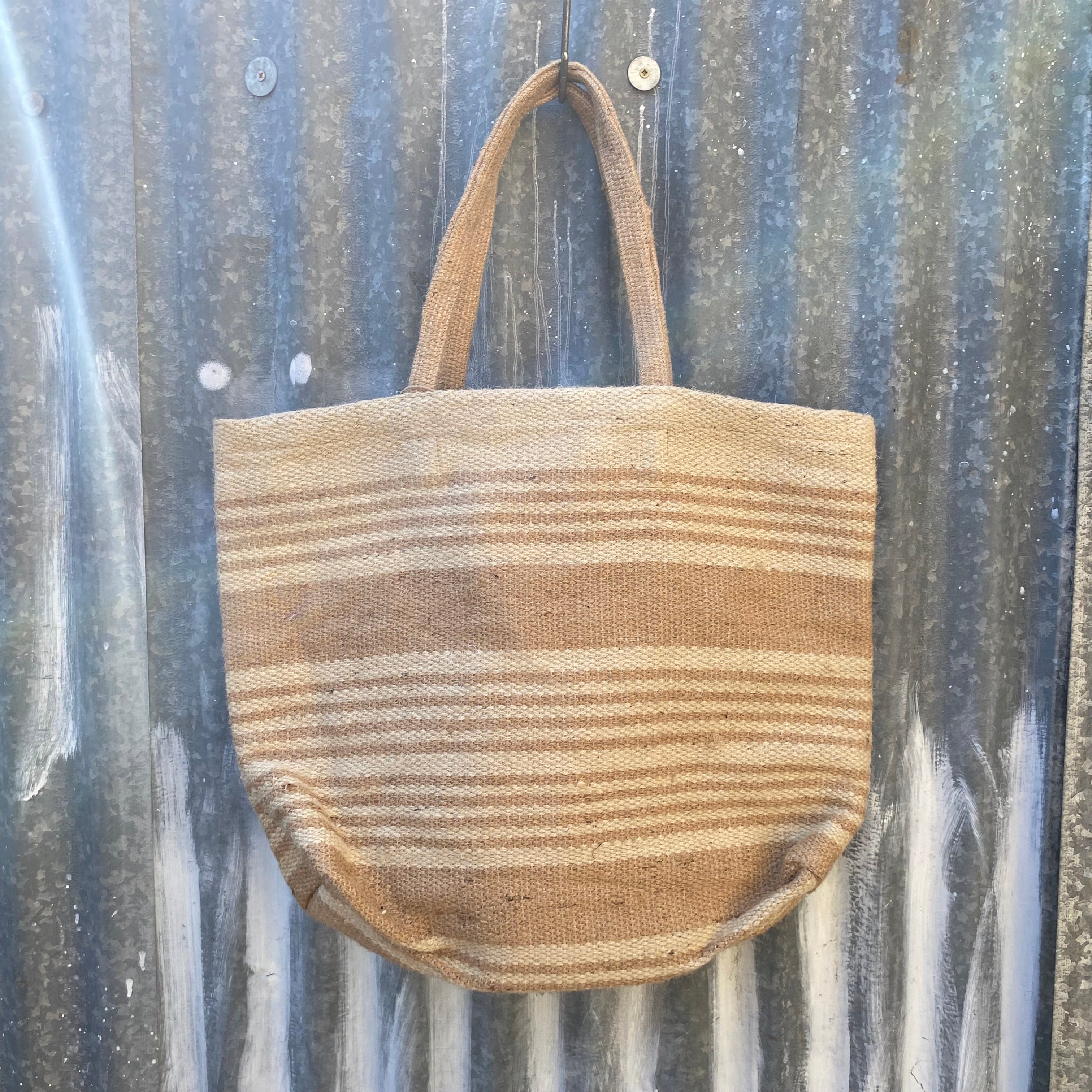 A woven jute tote bag with horizontal tan stripes hangs against a weathered corrugated metal wall.