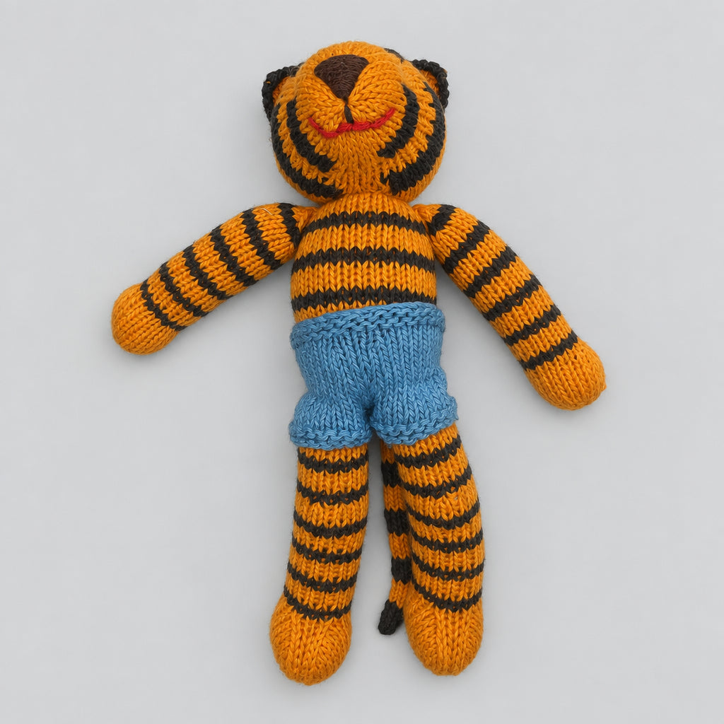 A handmade knitted tiger doll with orange and black stripes, wearing blue shorts, lying on a white fabric background.