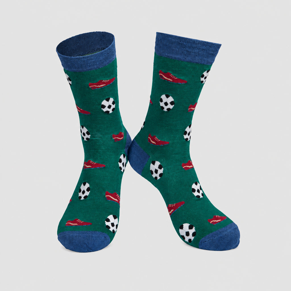 A pair of crew socks with a dark green background featuring a pattern of black and white soccer balls and red sneakers, with solid blue cuffs, heels, and toes, displayed on a grey background.