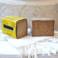 A bar of handmade Karwan soap displayed next to its yellow paper packaging box, resting on a white fringed textile against a white carved ceramic background.