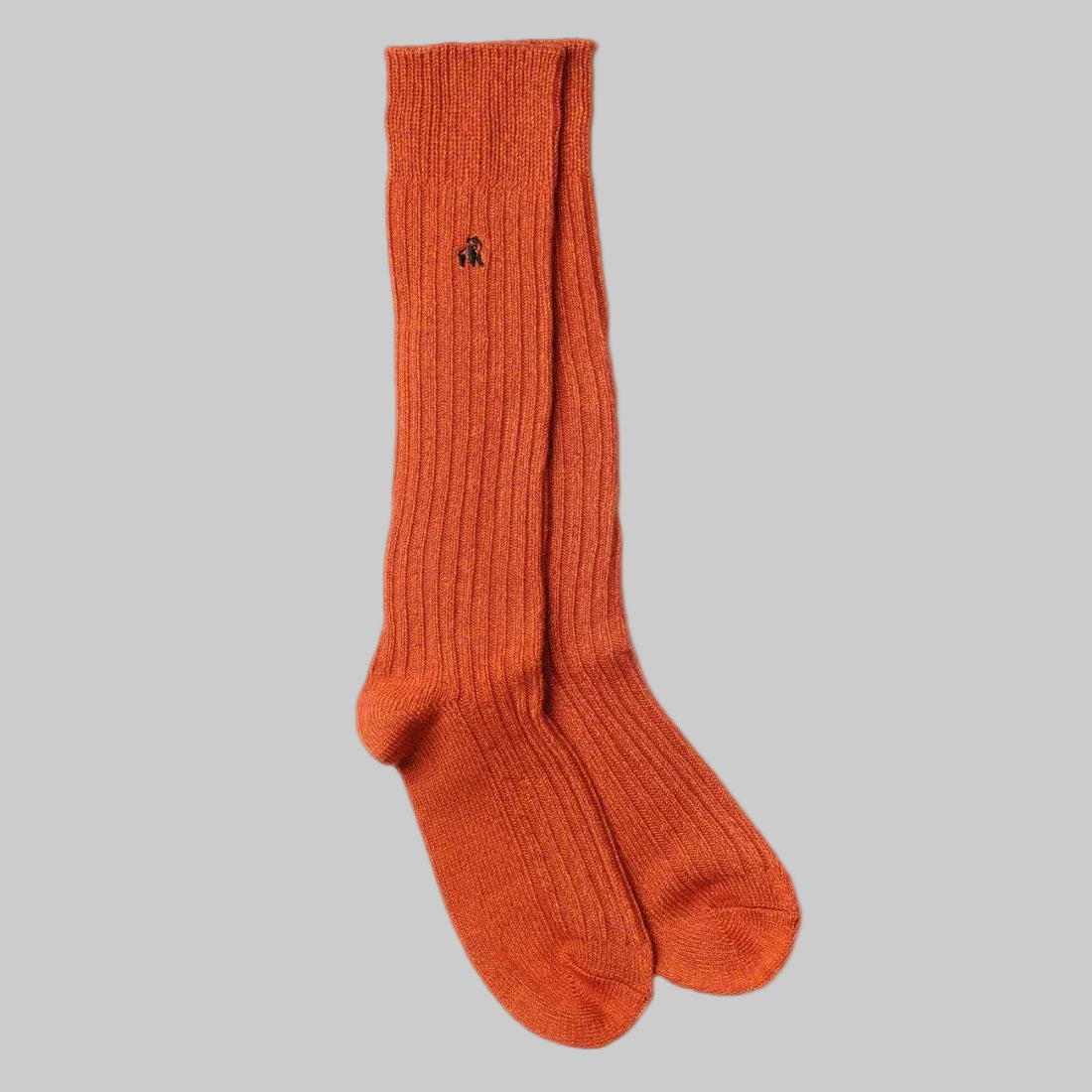 A pair of ribbed knit crew socks in a warm rust orange color, featuring a small black embroidered logo on the upper cuff, displayed against a solid white background.